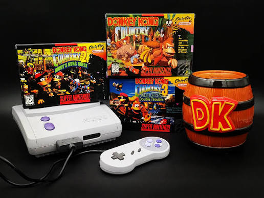 A Super Nintendo console and controller beside boxed copies of Donkey Kong Country 1, 2 and 3, with a DK barrel mug.